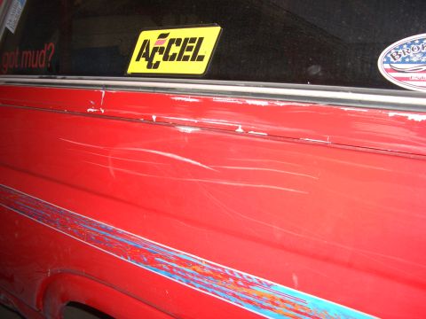 1985 Ford Bronco - A red vehicle's side panel shows significant scratches and a decorative stripe, with stickers on the rear window.