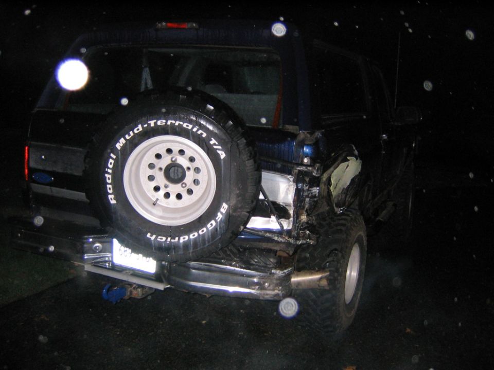 Rear of dark blue Ford Bronco at night with crushed right rear quarter and tailgate spare tire