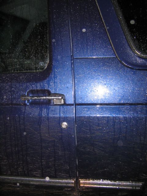 1994 Ford Bronco - A close-up view of a blue SUV's side door, covered in water droplets, with a visible logo.