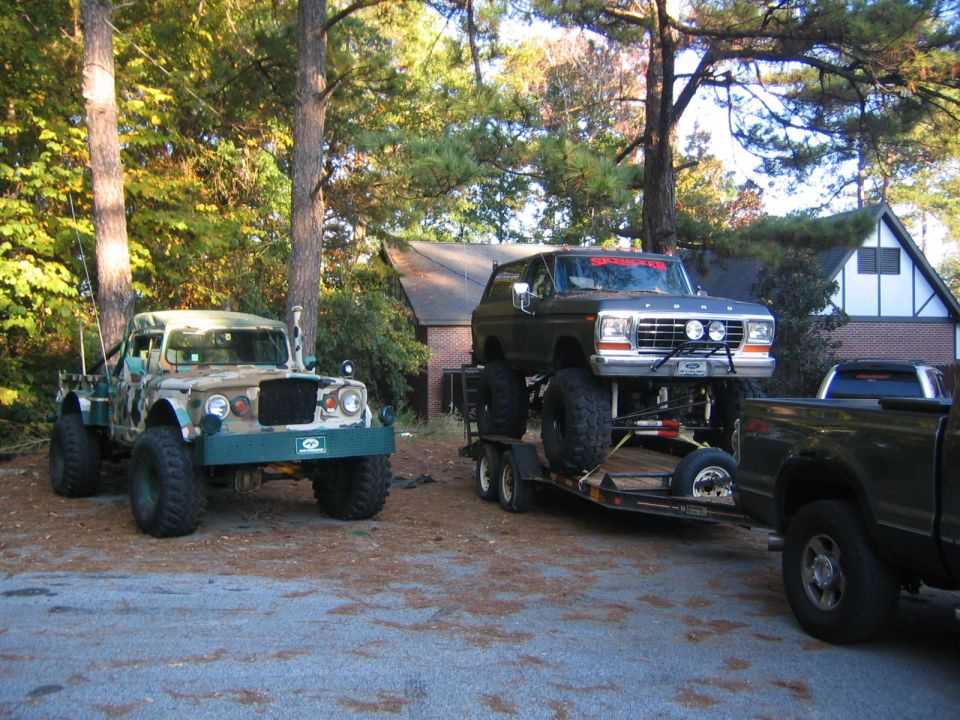 Lifted 1978 Ford Bronco on trailer, red text on windshield. Camouflage Jeep beside it
