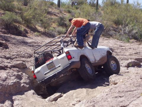 1993 Ford Bronco - Two people attempt to free a white off-road vehicle that is stuck on a rocky incline.