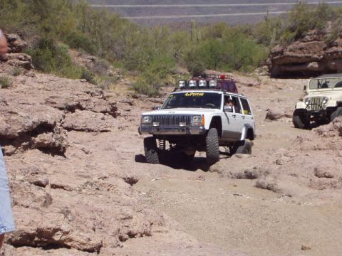 1993 Ford Bronco - A white Jeep Cherokee navigates a rocky desert trail, with another Jeep visible in the background.