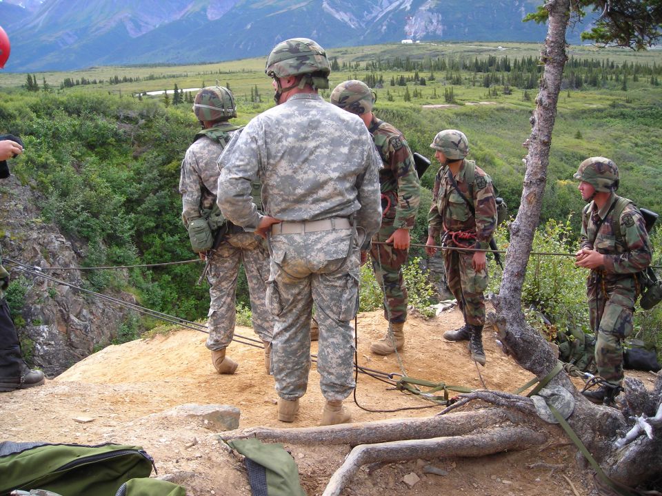 Military personnel preparing to rappel down a rocky slope with mountains in the background