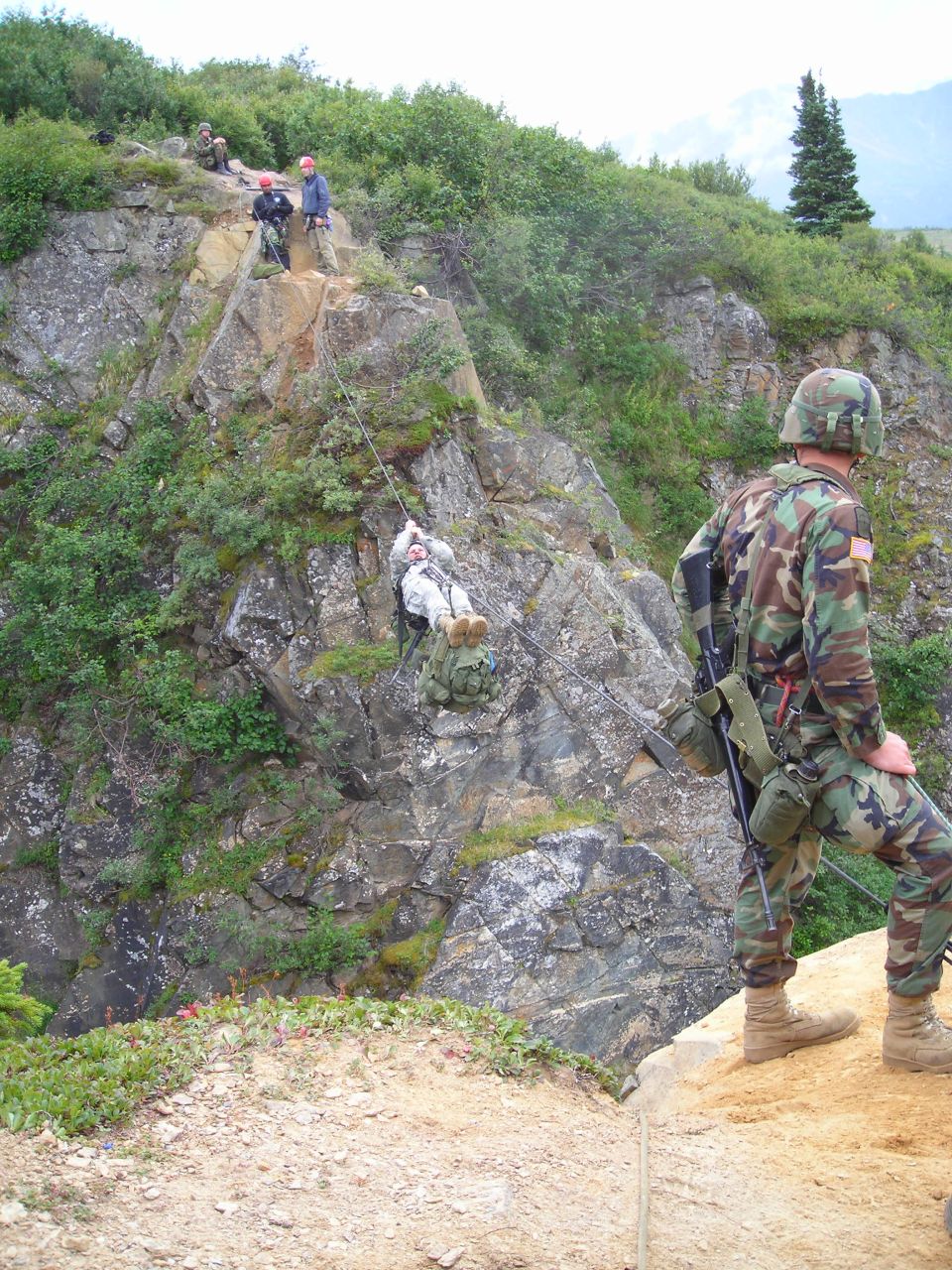 Military personnel rappelling down a rocky cliff face with a soldier standing guard