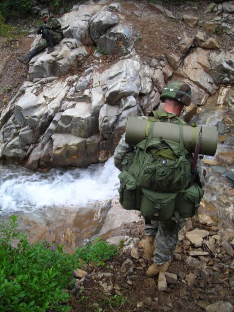 2003 Jeep TJ - Two soldiers equipped with heavy backpacks traverse a , rocky area alongside a flowing stream.