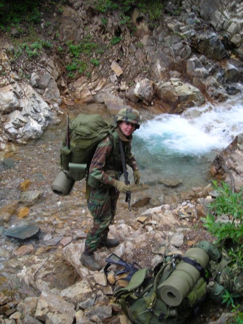 2003 Jeep TJ - A soldier equipped for a trek pauses by a flowing stream in a rocky, wooded area.