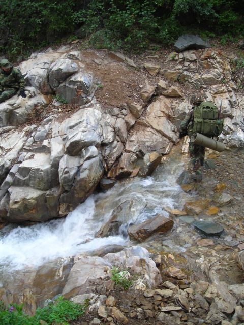 2003 Jeep TJ - Two soldiers navigate a challenging stream crossing in a rocky, wooded environment.