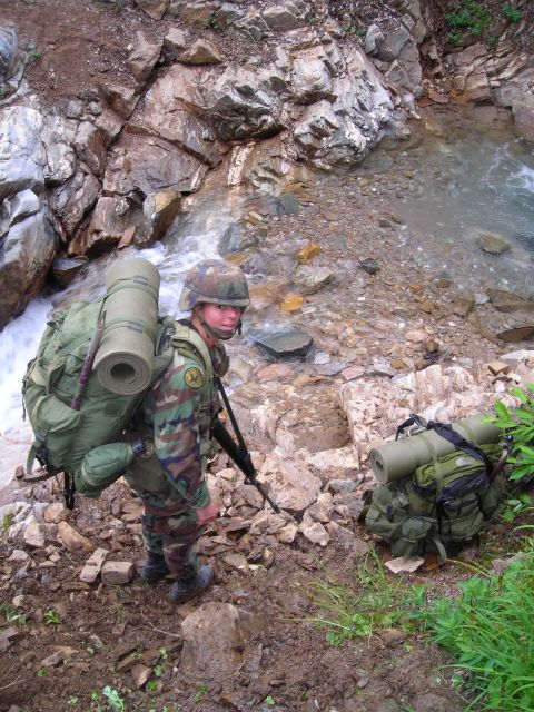 2003 Jeep TJ - A soldier equipped for a trek navigates a rocky terrain beside a flowing stream.