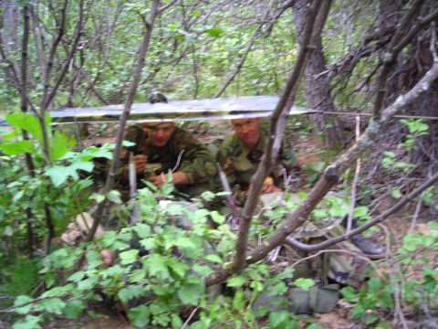 2003 Jeep TJ - Two soldiers in camouflage uniforms are concealed in a wooded area, with one holding an object and the other eating.