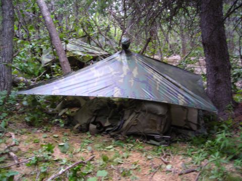 2003 Jeep TJ - A camouflage tarp is erected over backpacks in a wooded area, creating a makeshift shelter.