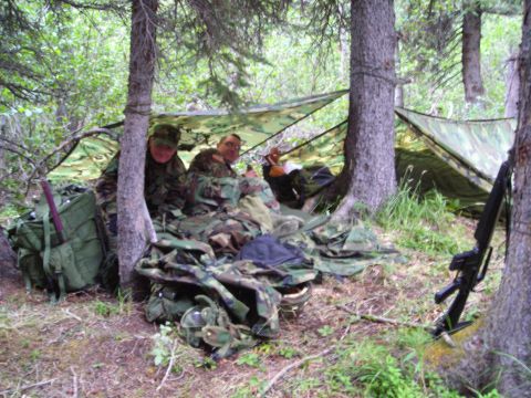 2003 Jeep TJ - Two individuals are sheltering under a camouflage tarp in a forest, with various pieces of military equipment around them.