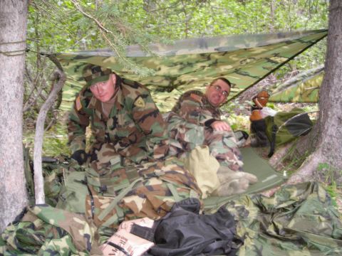 2003 Jeep TJ - Two soldiers rest under a camouflage tarp shelter amidst trees and military equipment in a wooded area.