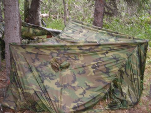 2003 Jeep TJ - A makeshift shelter made of camouflage fabric is erected amongst trees in a forest.