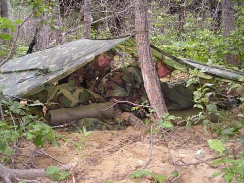 2003 Jeep TJ - Two individuals in military camouflage attire are sheltered beneath a tarp in a forest clearing.