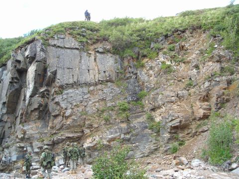 2003 Jeep TJ - A group of soldiers are positioned at the bottom of a steep, rocky incline with vegetation growing on top.