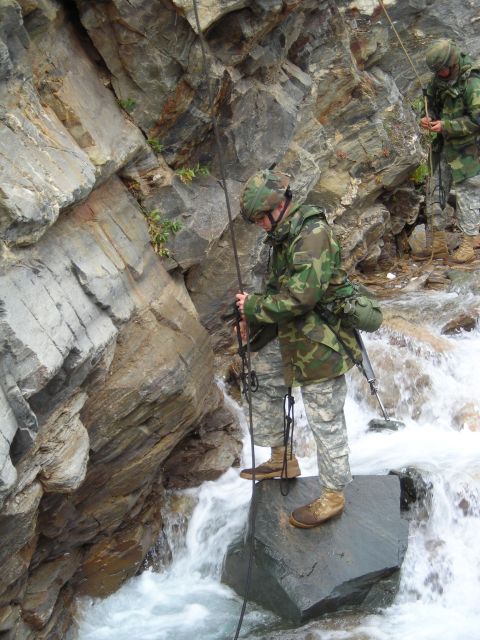 2003 Jeep TJ - Two soldiers in camouflage gear navigate a rocky stream, using ropes for assistance as they descend.