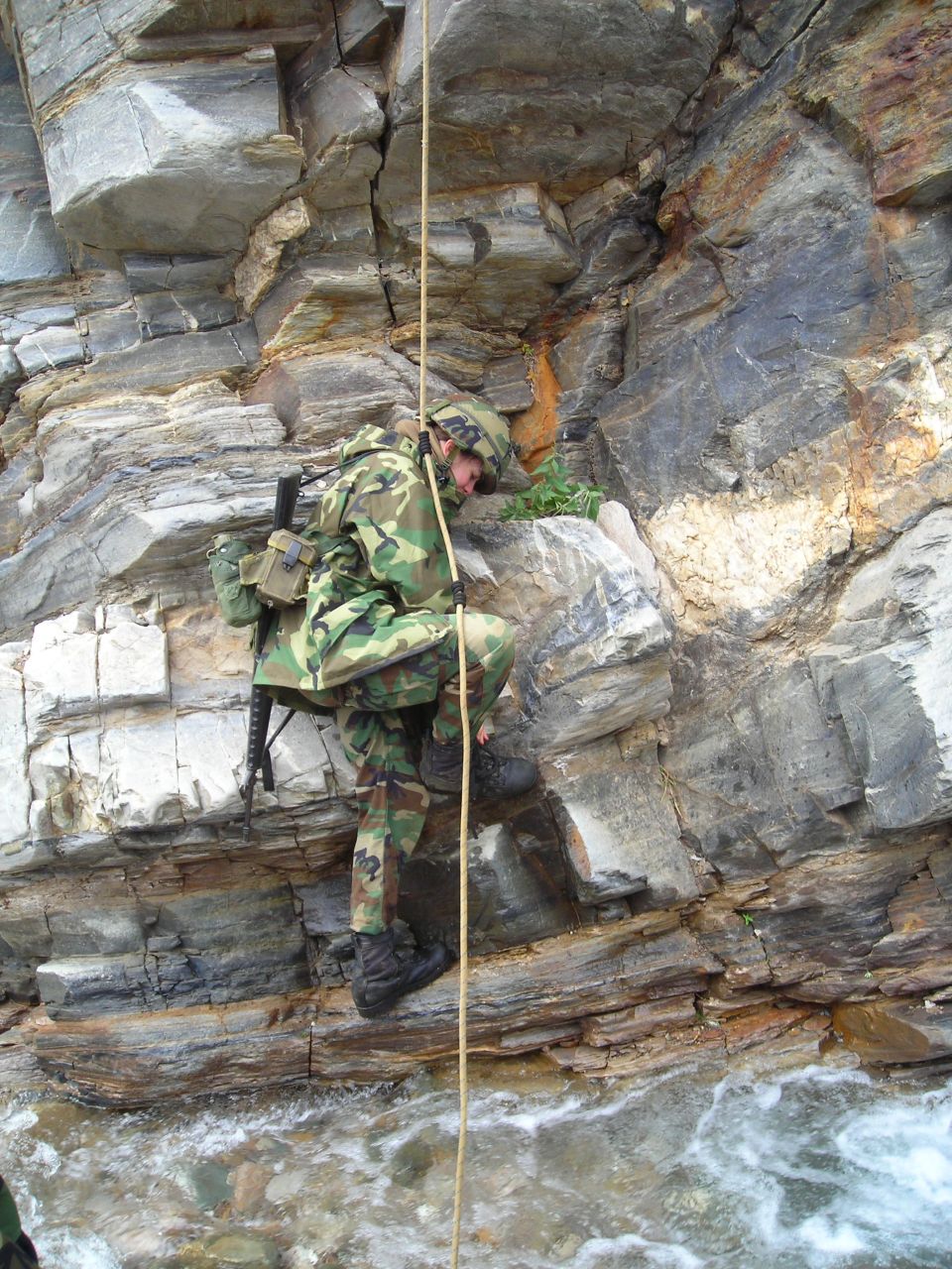 Soldier in camouflage rappelling down a rocky cliff with a rifle on their back, river below