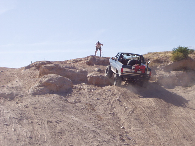 White roll-caged Ford Bronco climbing a dusty desert hill, rear view