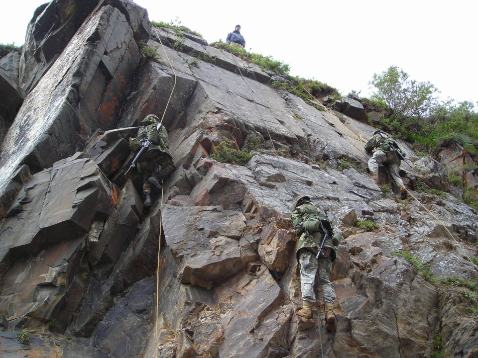 Soldiers in camouflage rappelling down a steep, rocky cliff face with ropes. One person watches from the top