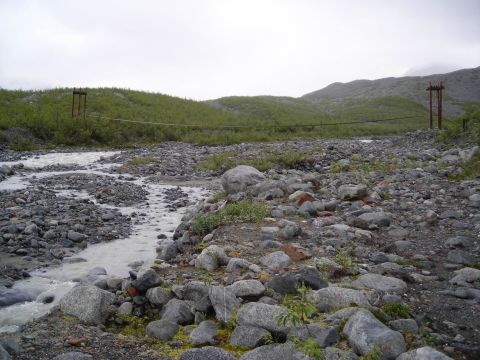 2003 Jeep TJ - A wide shot of a rocky riverbed with a suspension bridge spanning the water, set against a backdrop of green hills under a cloudy sky.