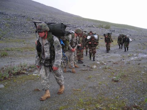 2003 Jeep TJ - A group of soldiers trek through a desolate, rocky landscape, carrying heavy packs on their backs.