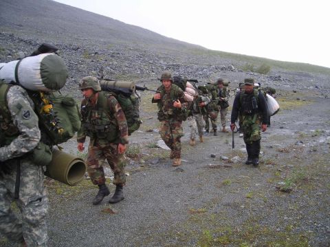 2003 Jeep TJ - A group of soldiers marches across a desolate, rocky terrain, each burdened with substantial backpacks and gear.