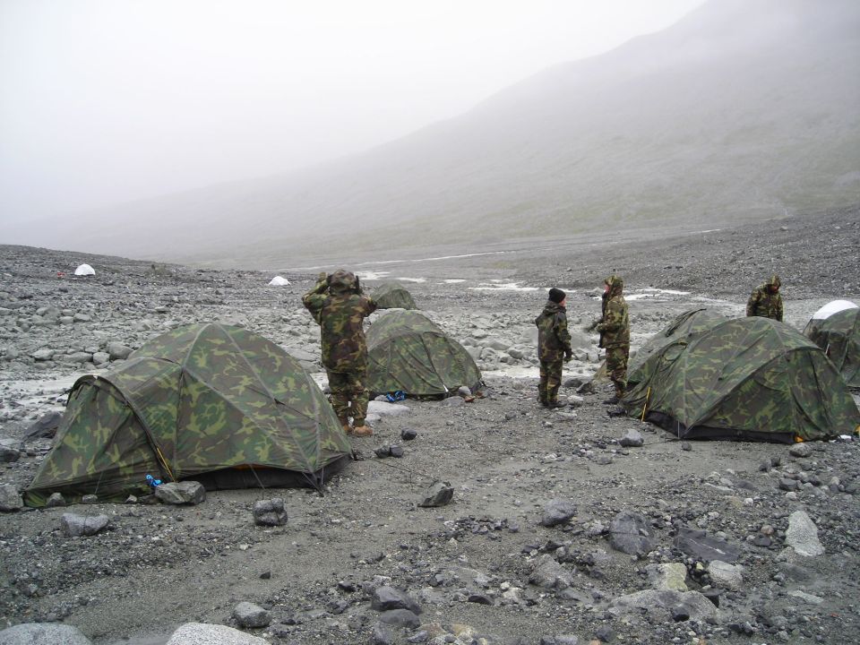 Several camouflage tents are set up on a rocky, barren ground with a foggy mountain in the background. People in camouflage uniforms are standing near the tents