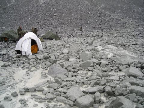 Several tents are set up on a rocky, barren slope with a small stream running through it. People are visible n