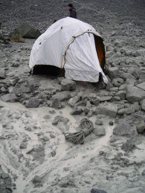 White tent pitched on rocky, muddy ground with a coiled rope in the foreground