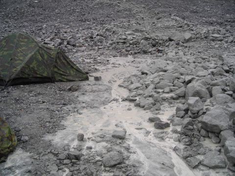 Camo tent pitched on rocky terrain with a muddy stream flowing nearby