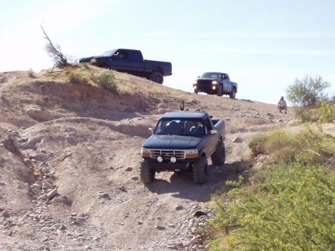 1993 Ford Bronco - A dark blue pickup truck navigates a steep, rocky incline with two other trucks visible in the background.