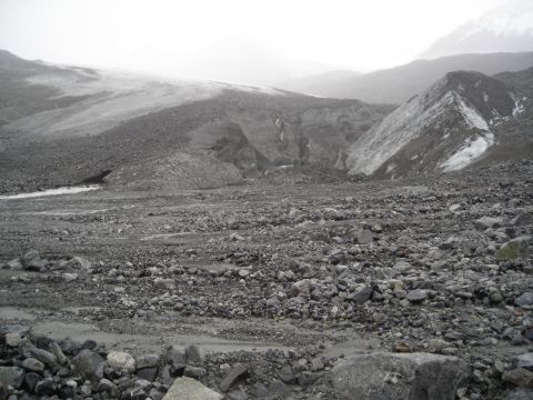 Rocky glacial moraine with a stream bed and distant glacier under a foggy sky