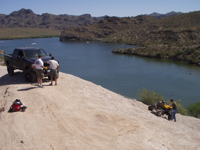 Lifted black pickup on a sandy desert bluff above a lake with a quad below