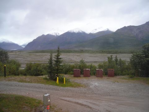Mountain range with snow-capped peaks under a cloudy sky, overlooking a wide, gravelly valley with trees and f