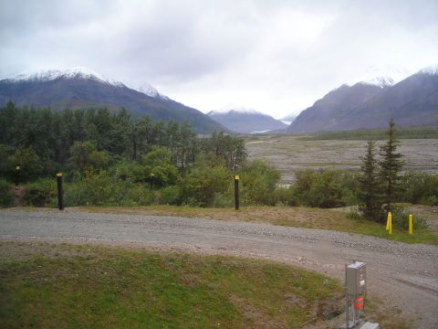 Mountain range with snow-capped peaks, trees, and a gravel road with RV hookups in the foreground