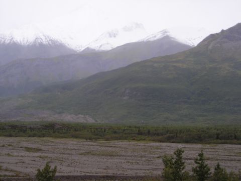 Snow-capped mountains rise above a green, forested valley and a gravelly riverbed