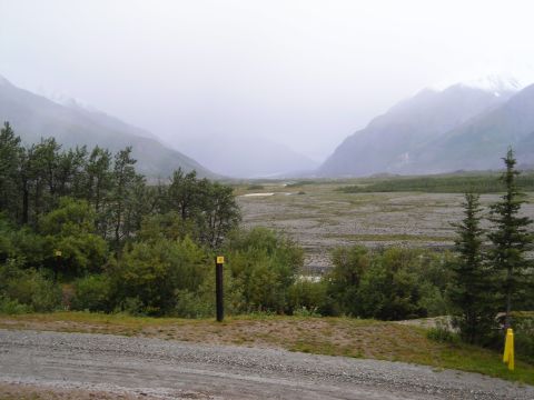 Mountain valley with trees and a gravelly riverbed under a foggy sky. Two yellow markers are visible
