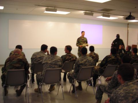 Military personnel seated in chairs facing a whiteboard and two standing figures, one presenting