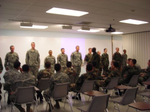 Group of soldiers standing and sitting in a room with a projector screen