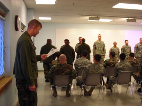 Military personnel standing and seated in a room, one reading a document