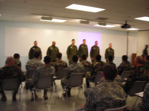 Six service members standing at the front of a briefing room facing a seated audience in camouflage uniforms