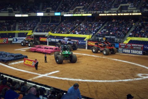 1987 Ford Bronco - Monster trucks are prepared for an event in a stadium, with multiple vehicles and a crowd watching.