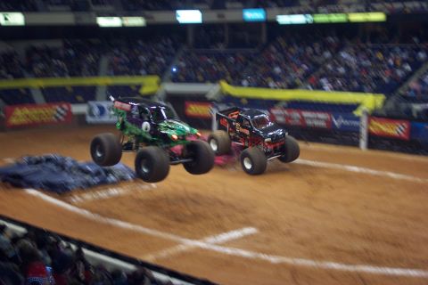 1987 Ford Bronco - Monster trucks compete in an arena, with two vehicles captured mid-air during a jump.