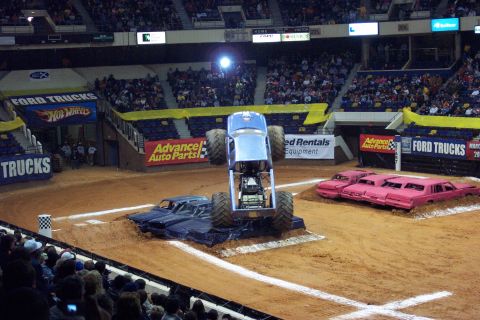 1987 Ford Bronco - A monster truck performs a stunt, jumping over a row of cars during an event.