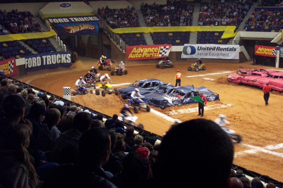 Crushed cars and ATVs on an arena dirt floor with a crowd watching