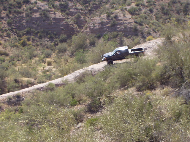 Dark Ford Bronco climbing a steep rock slab on a brushy desert hillside, seen from a distance