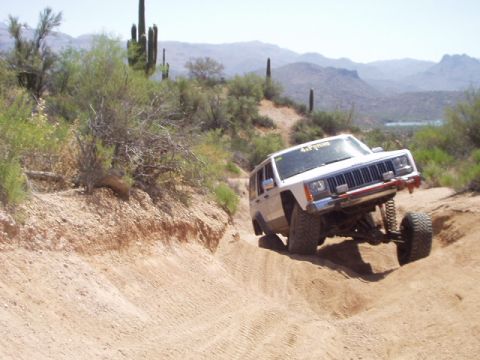 1993 Ford Bronco - A white Jeep Cherokee XJ navigates a steep, sandy off-road trail with cacti and mountains in the background.