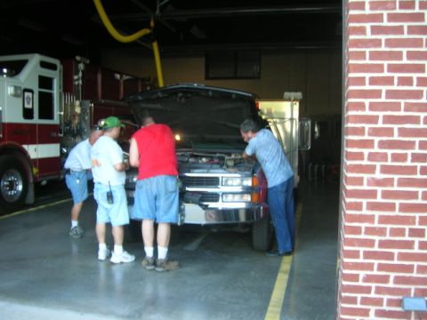 1989 Ford Bronco - Four men gather around the open hood of a white pickup truck parked inside a fire station bay, with a red-and-white fire engine to the left…