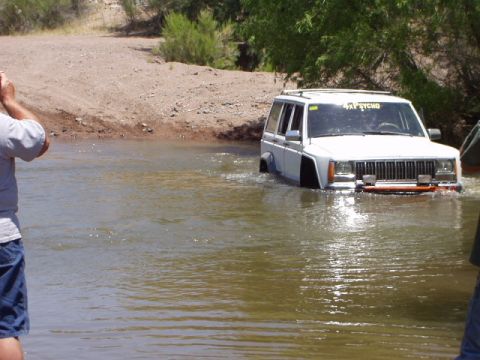 1993 Ford Bronco - A white Jeep Cherokee navigates through a body of water, displaying a '4x4 PSYCHO' sticker on its windshield.