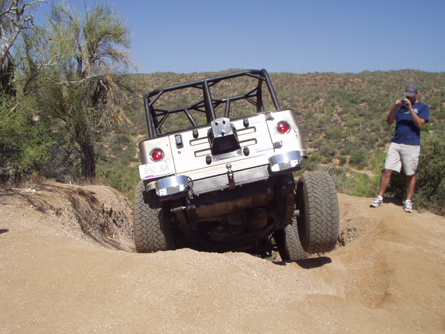 White caged topless Ford Bronco trail rig flexing over a dirt berm, rear view